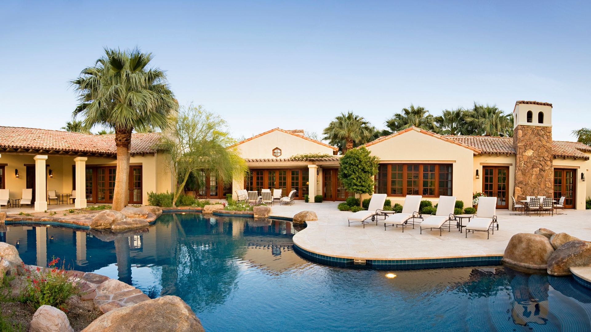 Two-story beige house with arched window, palm trees, and green lawn on a sunny day.