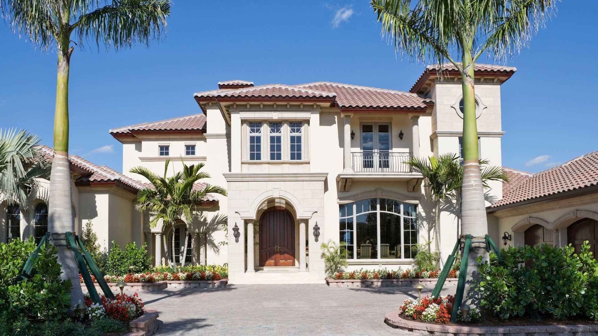 Beige mansion with red-tiled roof, palm trees, and brick driveway under a blue sky.