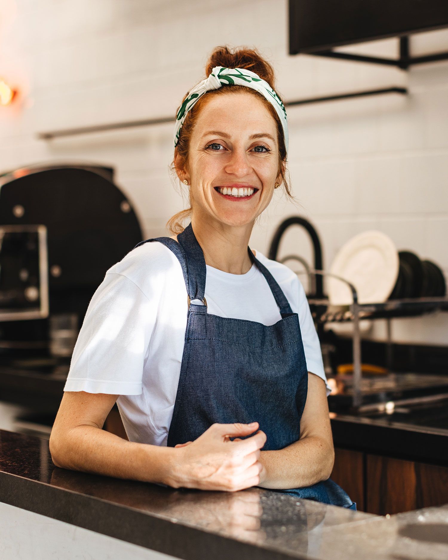A woman in an apron is smiling while sitting at a counter in a kitchen.