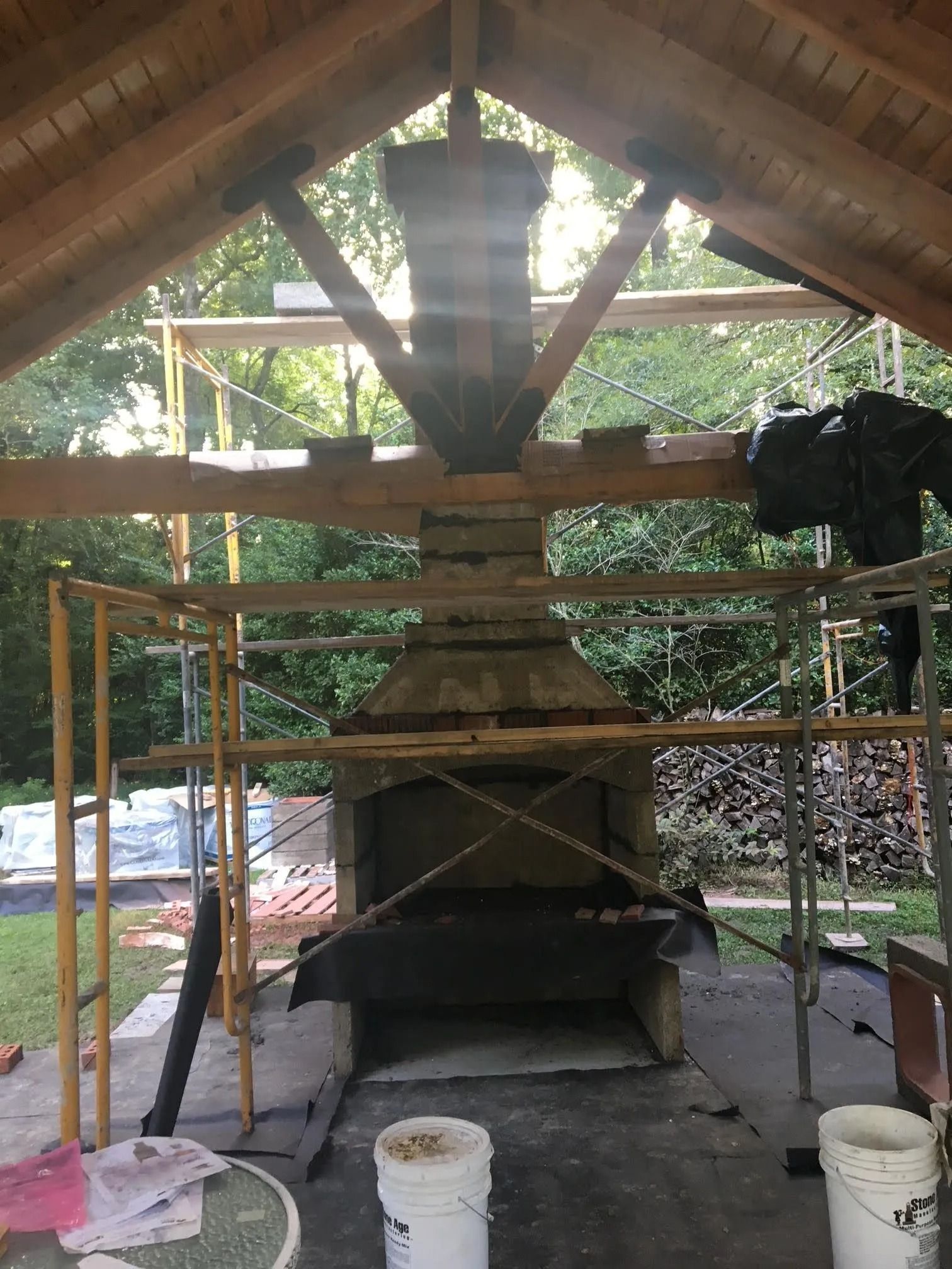Outdoor brick fireplace under a wooden pavilion, with scaffolding and buckets around it