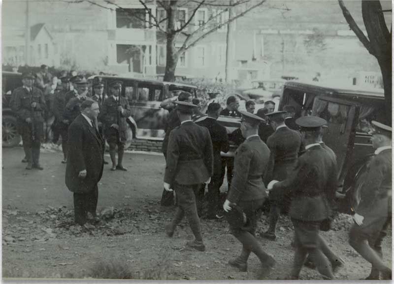 Group of uniformed men walking alongside a civilian man near parked cars and buildings, possibly outdoors.