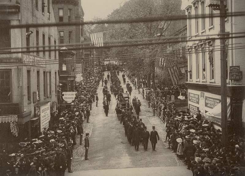 Parade marching down a street lined with buildings and spectators, American flags hanging.