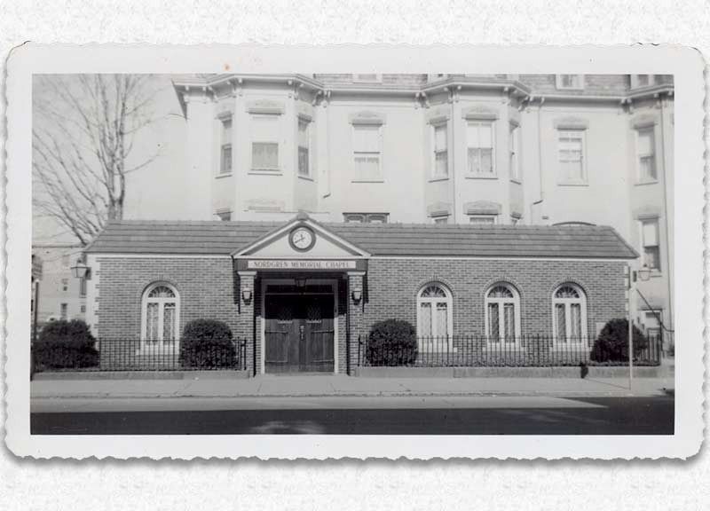Brick building with arched windows and a sign, in front of a larger building with bay windows.