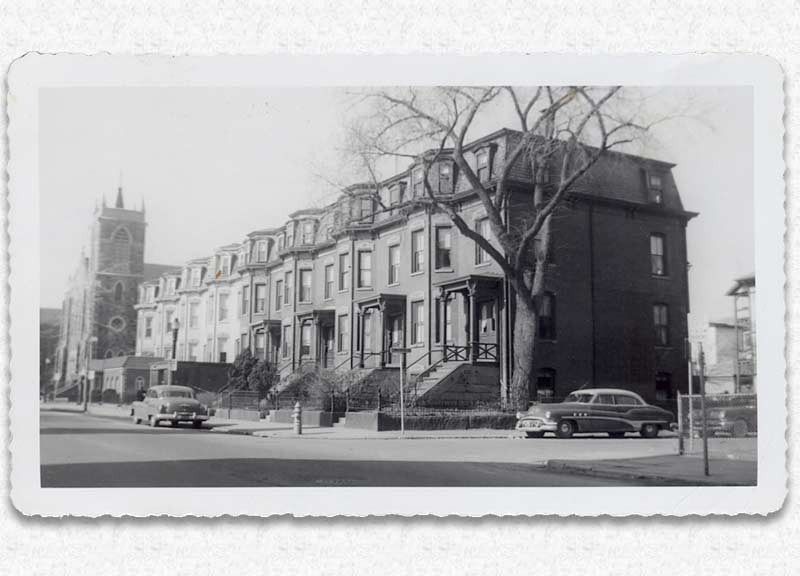 Row houses with ornate facades and cars on a city street; a church steeple is in the background.