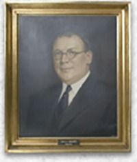 Portrait of a man in a suit and glasses, within a gold-colored frame, against a dark background.