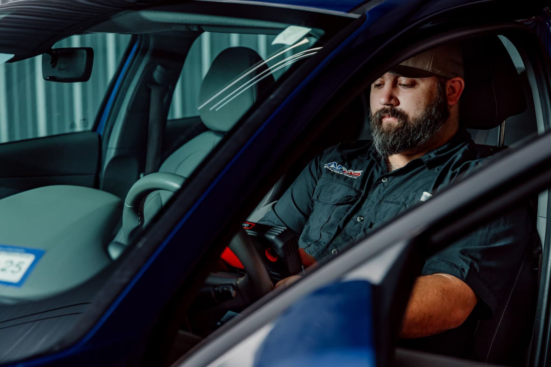 Man with beard sitting in the driver's seat of a blue car, looking down.