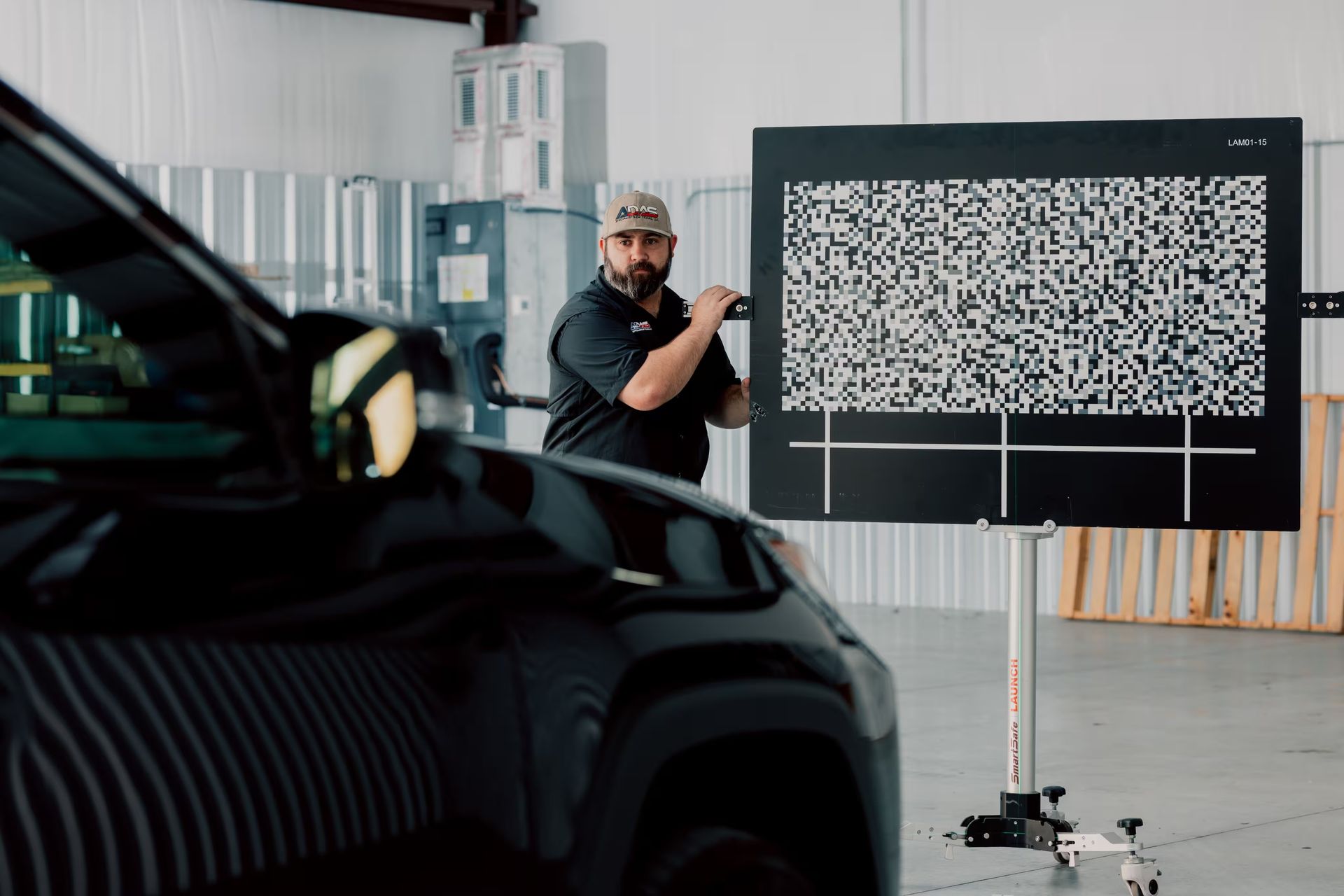 Mechanic using a device to inspect a black vehicle in a garage, next to a pattern board.