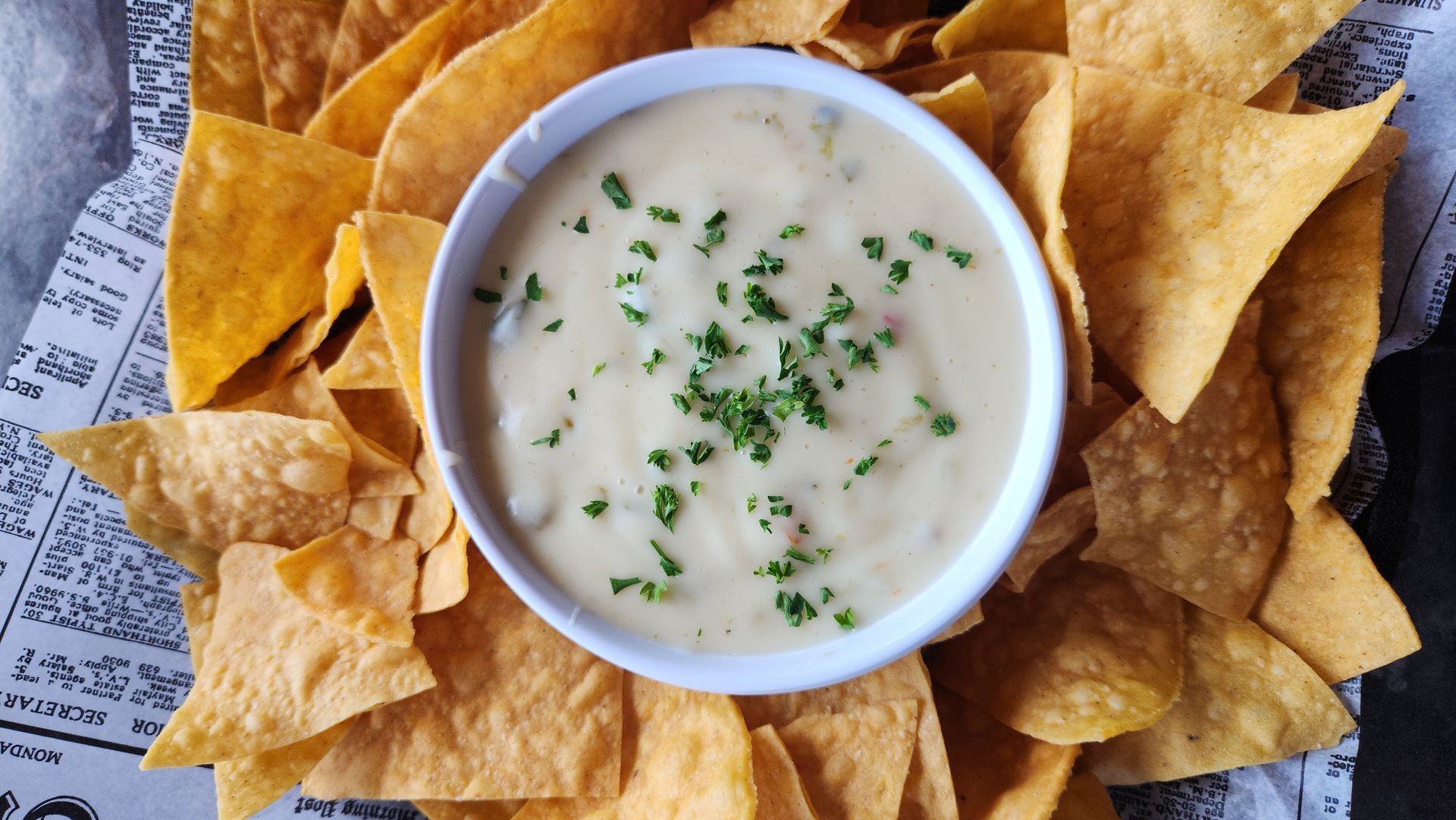 Bowl of queso surrounded by tortilla chips, garnished with chopped herbs.