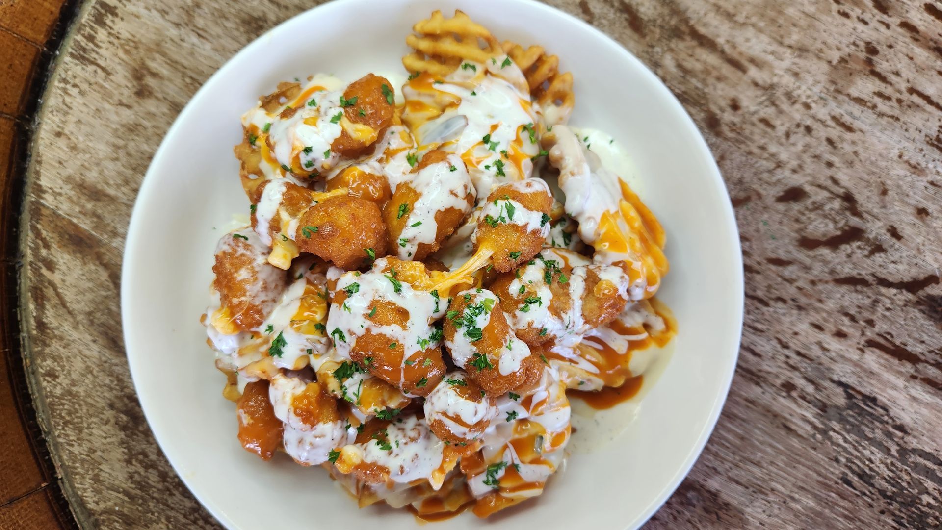 Close-up of a white bowl with waffle fries and orange-colored fried food, topped with white sauce and parsley.