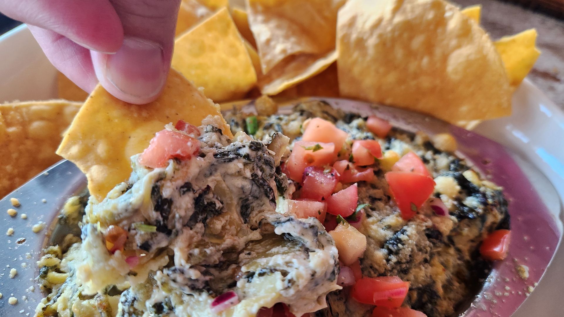 Hand dipping a tortilla chip into spinach and artichoke dip, topped with tomatoes and served with chips.