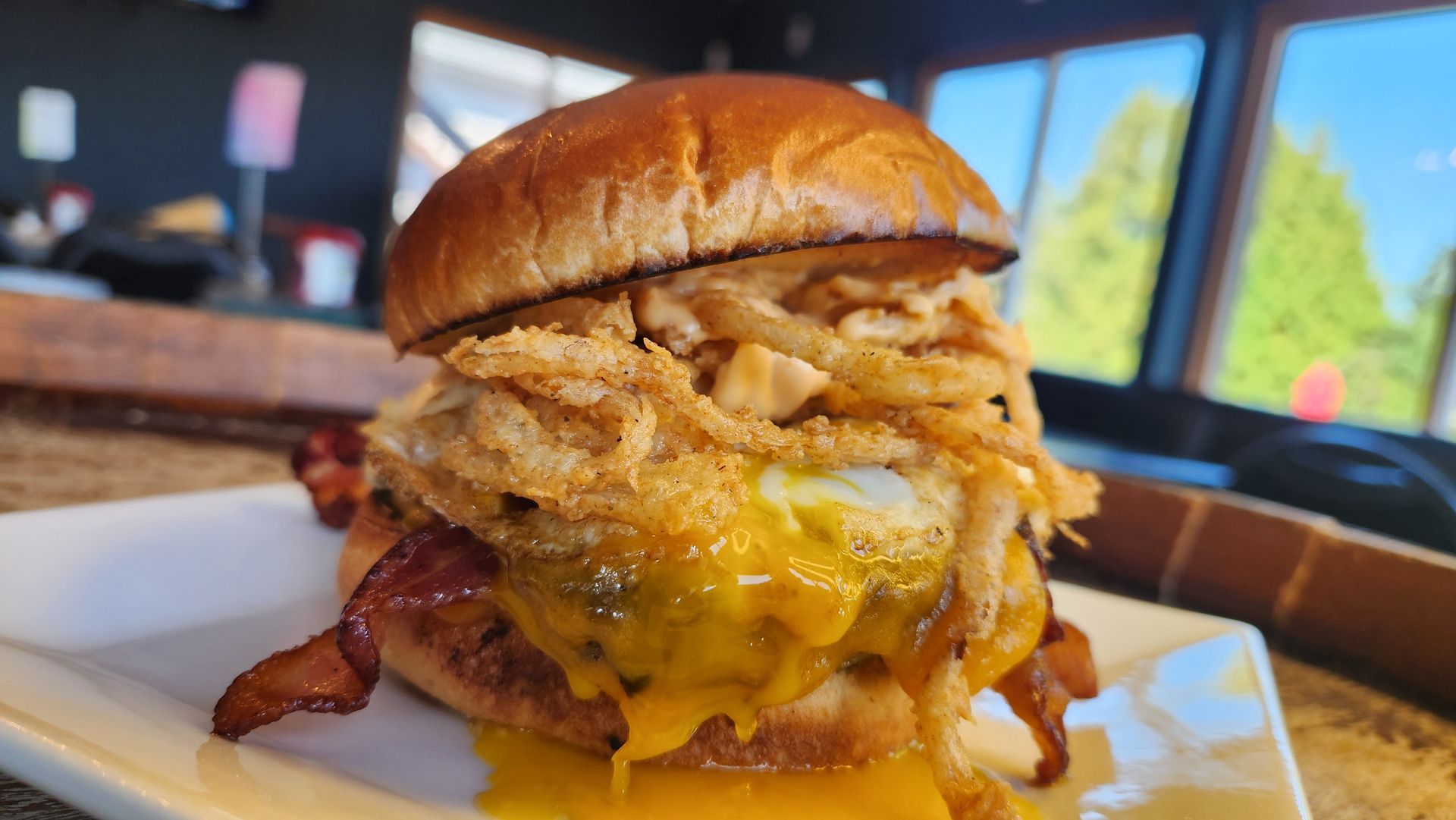 Burger on white plate, loaded with bacon, fried onions, cheese, and egg; near window.