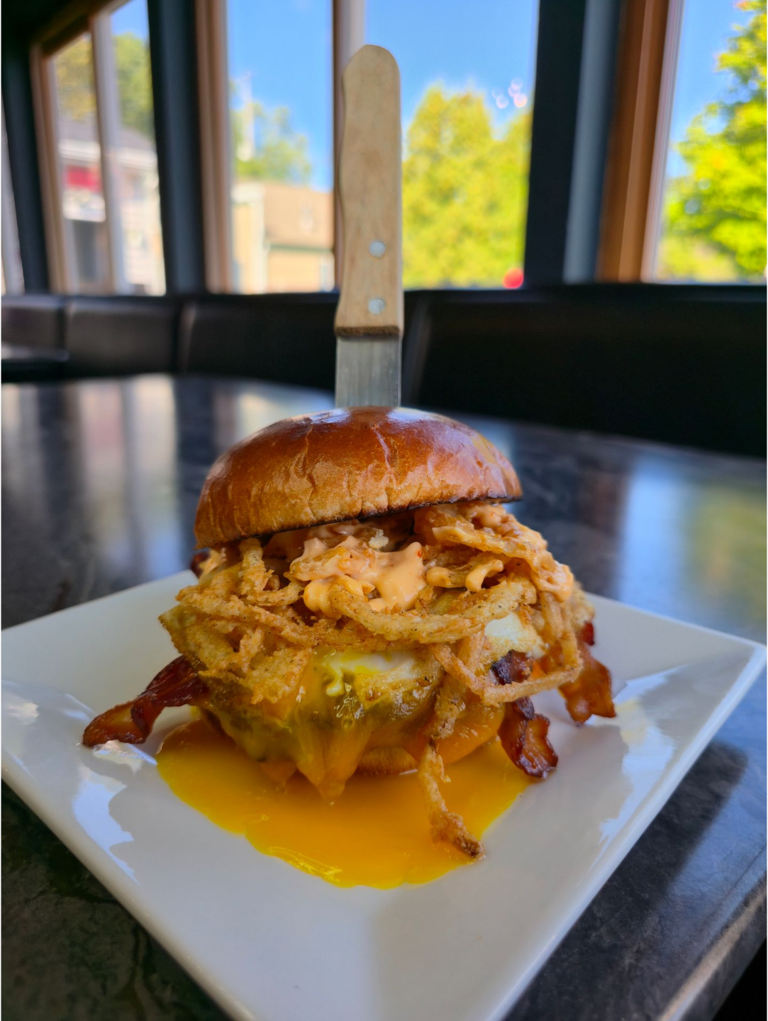 Burger with fried egg, bacon, and onion rings on a white square plate, held together with a knife.
