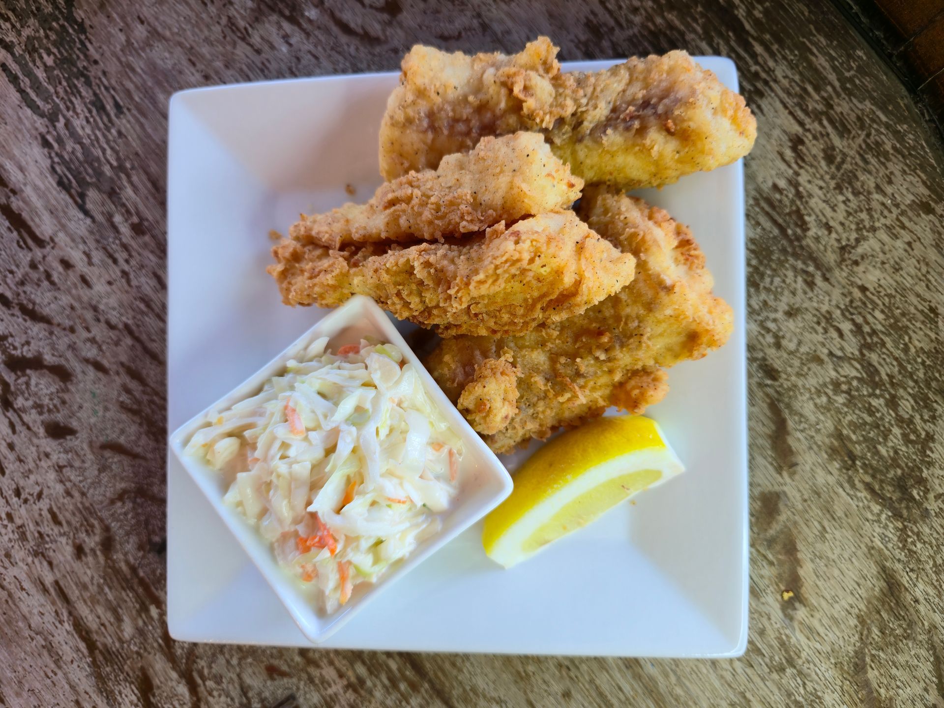 Fried fish, coleslaw, and lemon wedge on a white square plate, set on a wooden surface.