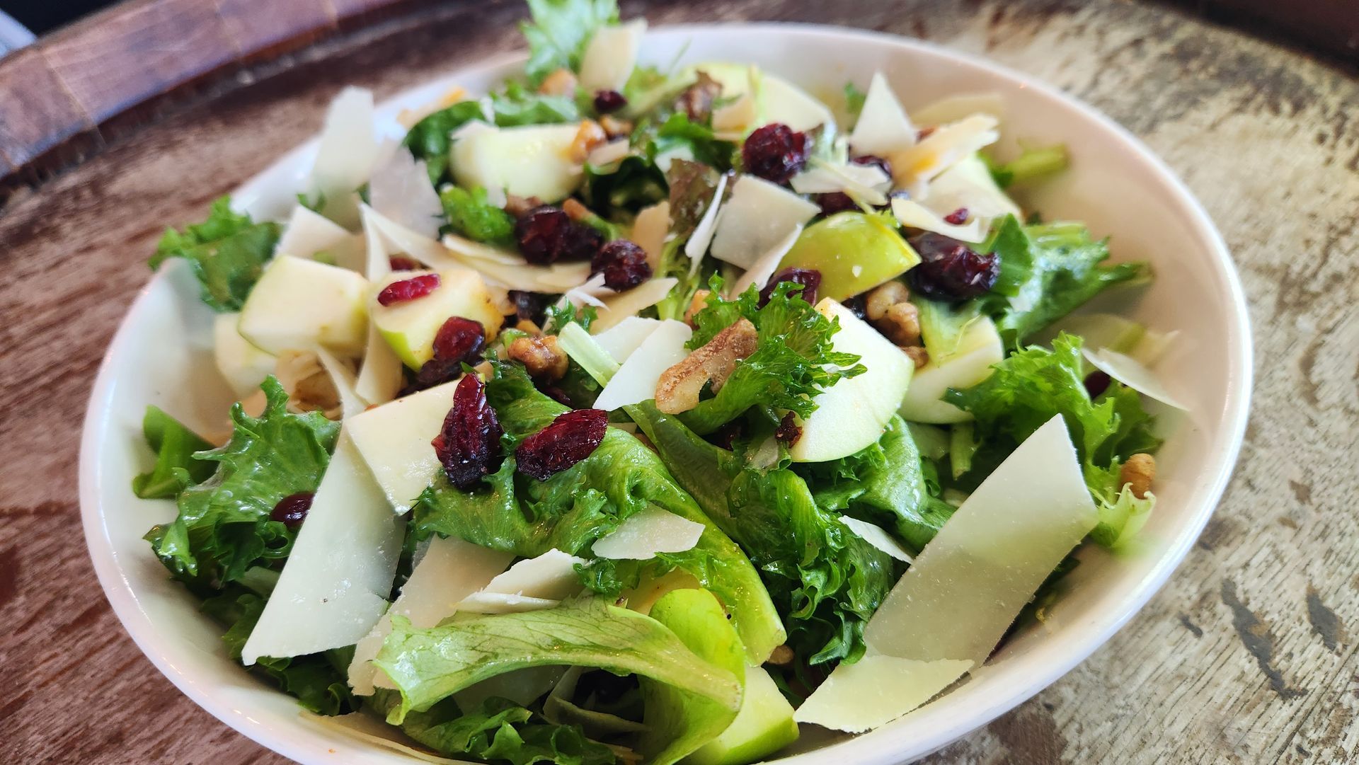Salad with greens, apple slices, cranberries, walnuts, and shaved cheese in a white bowl.
