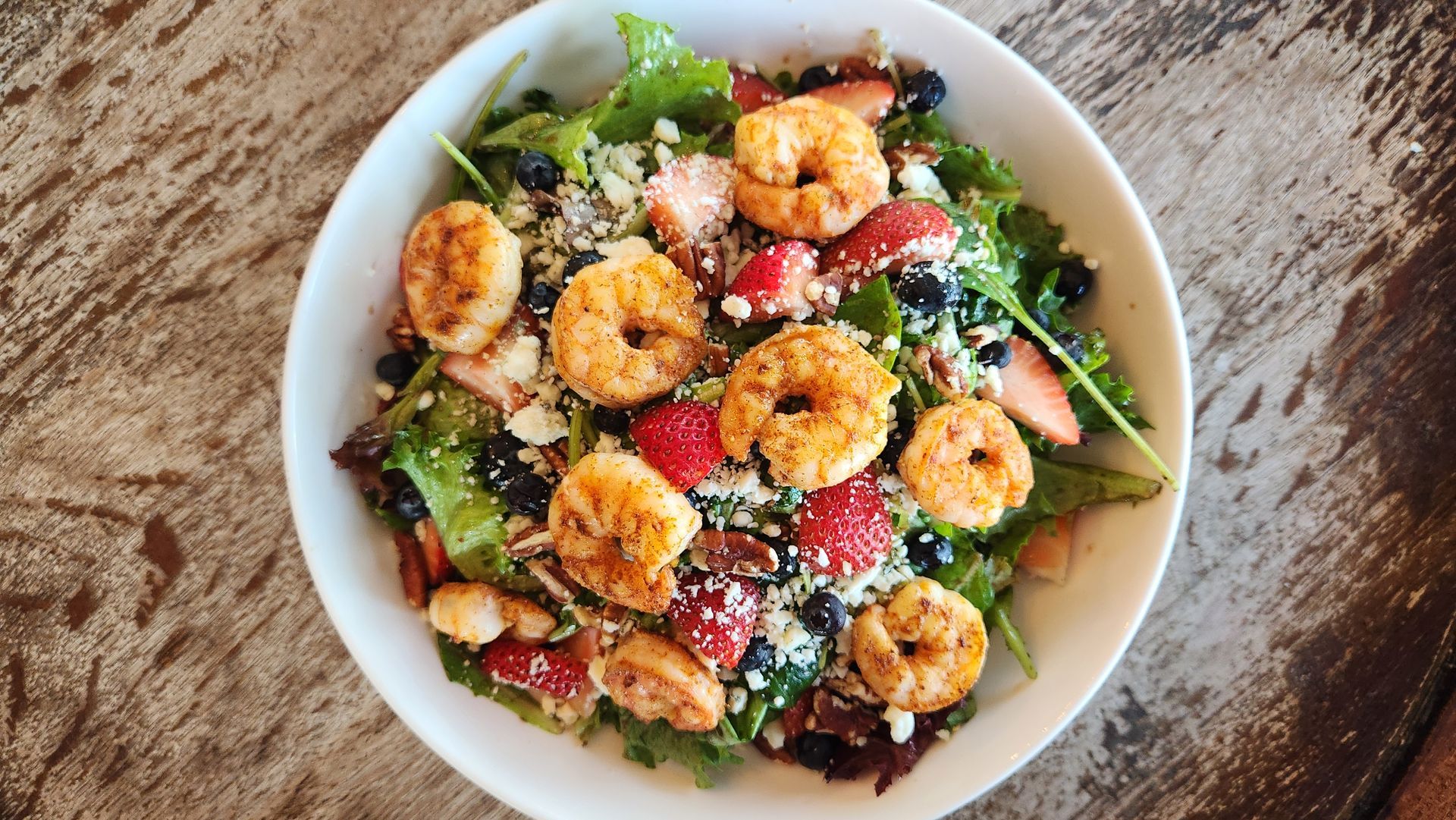Shrimp salad with strawberries, blueberries, and nuts in a white bowl on a wooden table.