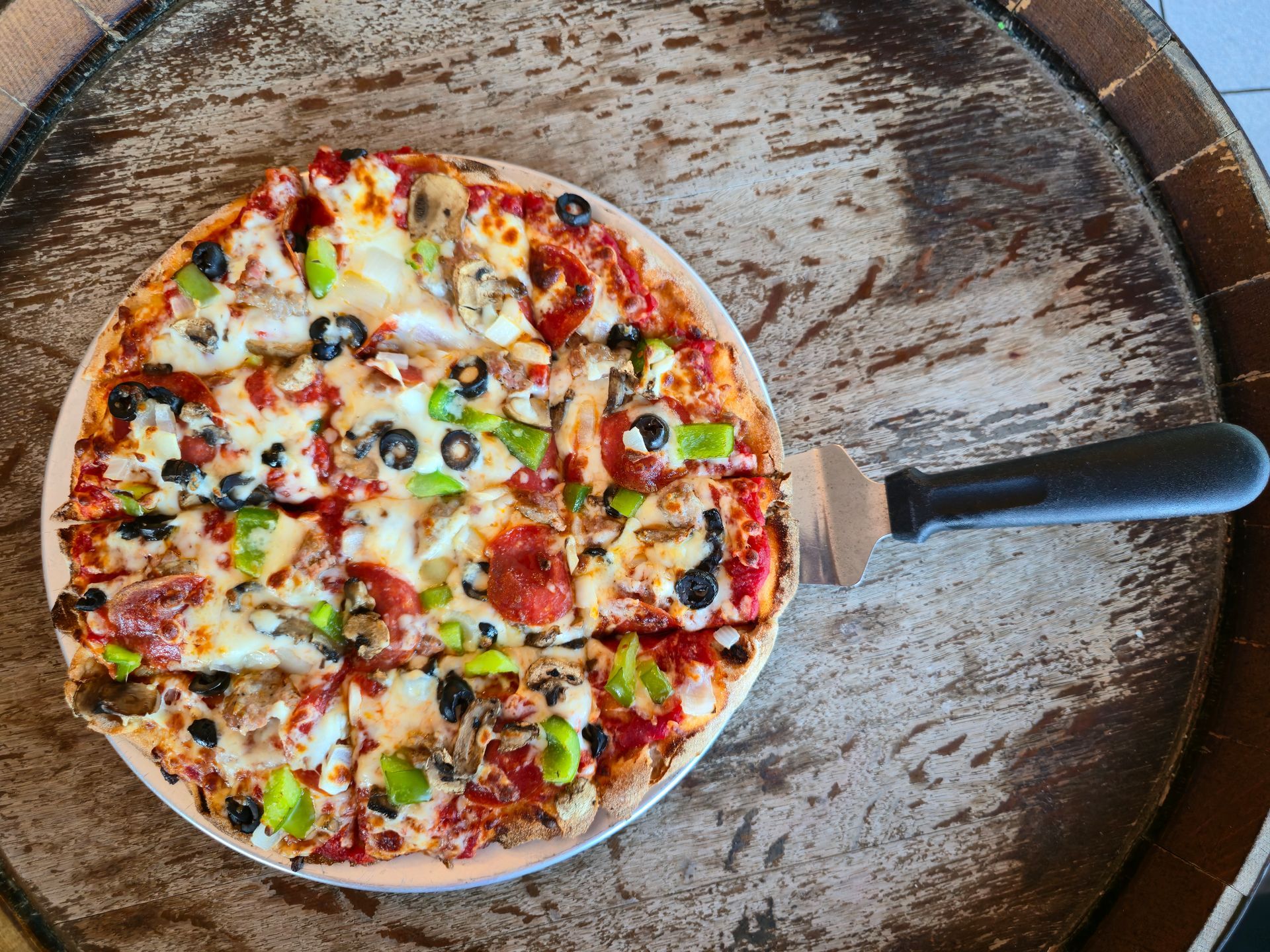 Pizza with toppings on a plate, next to a pizza server, sits on a wooden barrel.