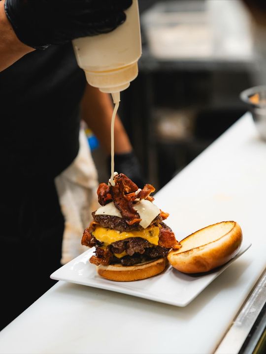 Chef topping a large burger with sauce; stacked patties, cheese, bacon, and bun.