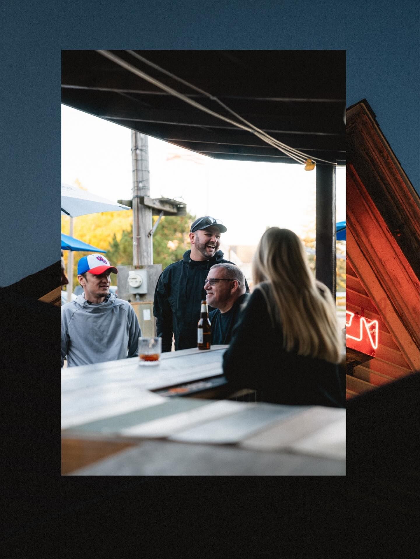 Four people at a table, under a wooden roof. Sunlight, mountains in the background. Two men and woman, smiling.