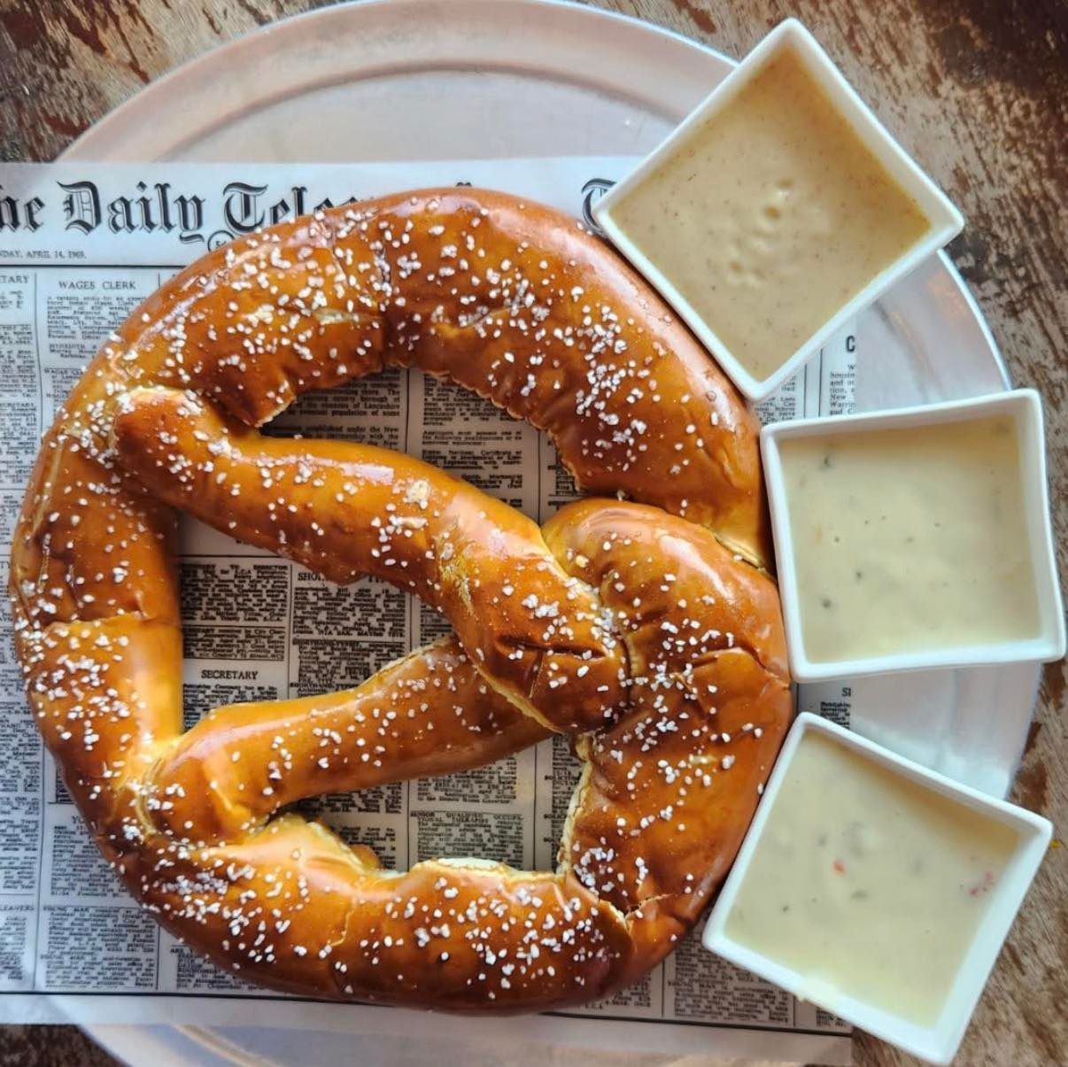 Giant pretzel with three dipping sauces on a newspaper-lined plate.