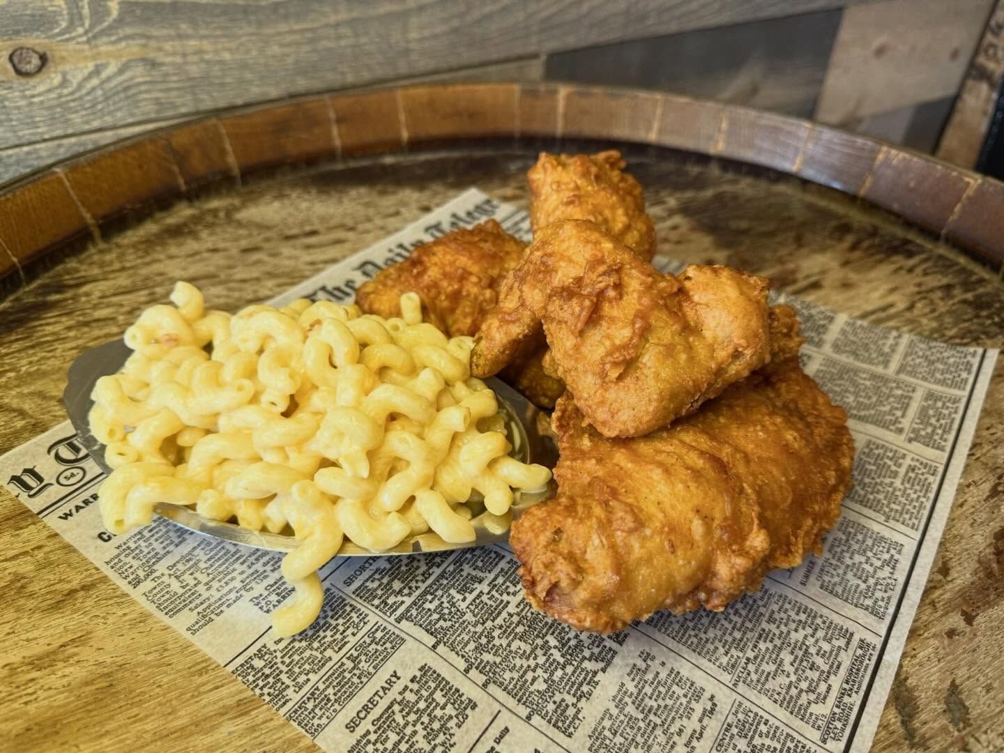 Macaroni and cheese with fried chicken on a newspaper-lined plate, atop a wood surface.