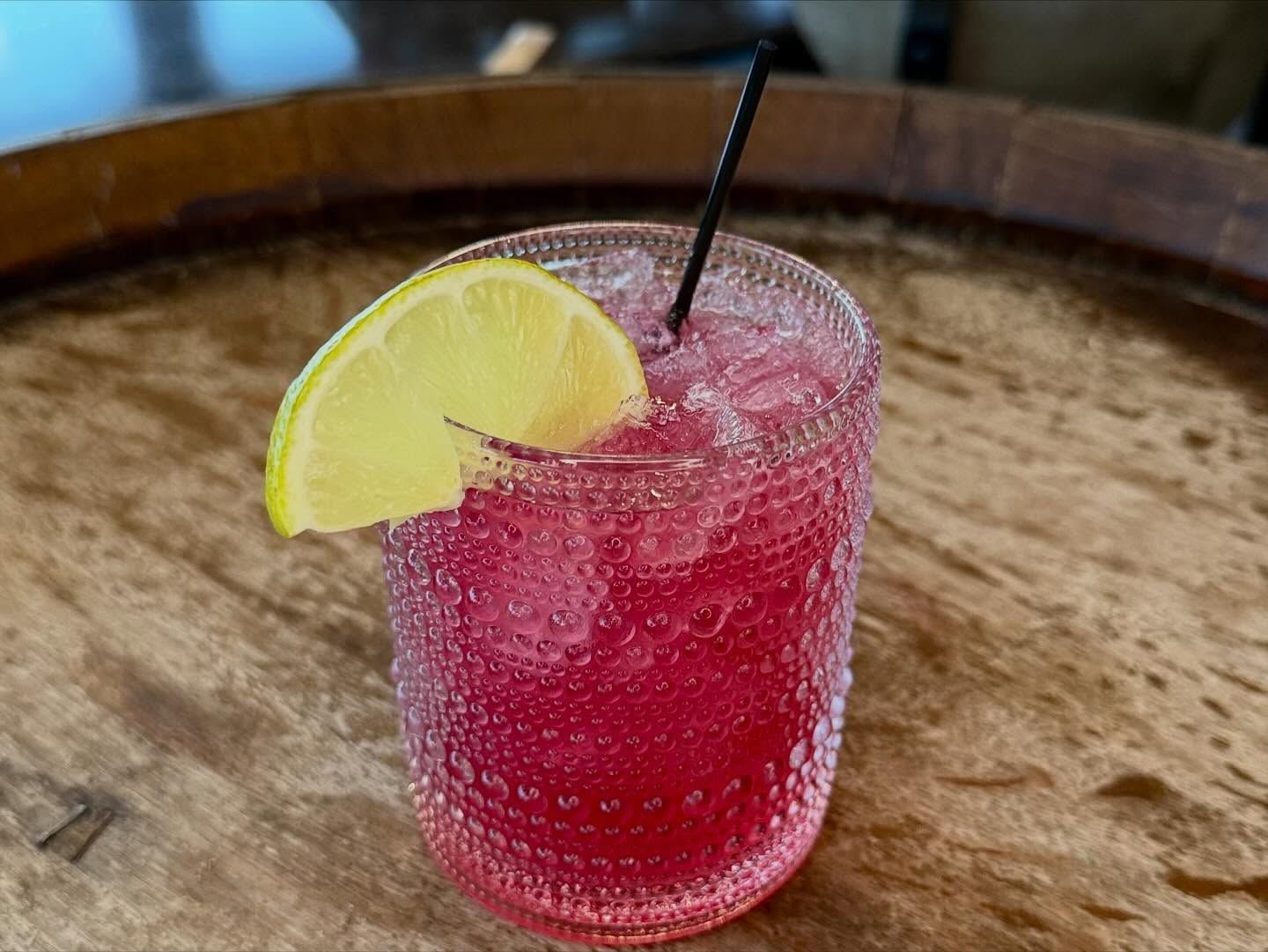 A vibrant pink cocktail in a textured glass, garnished with a lime slice and a black straw, on a wooden surface.