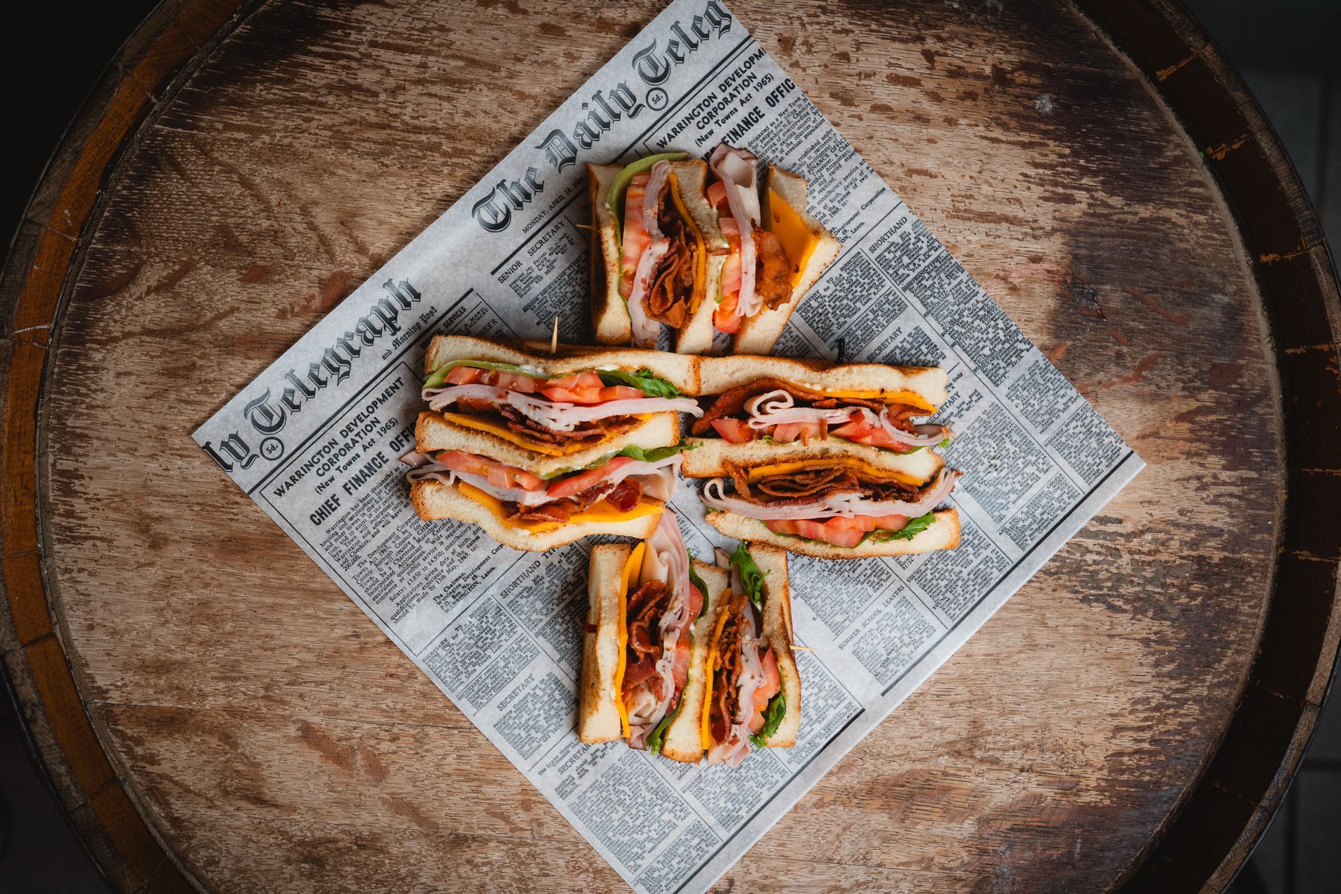 Club sandwiches on a newspaper, arranged on a wooden barrel.