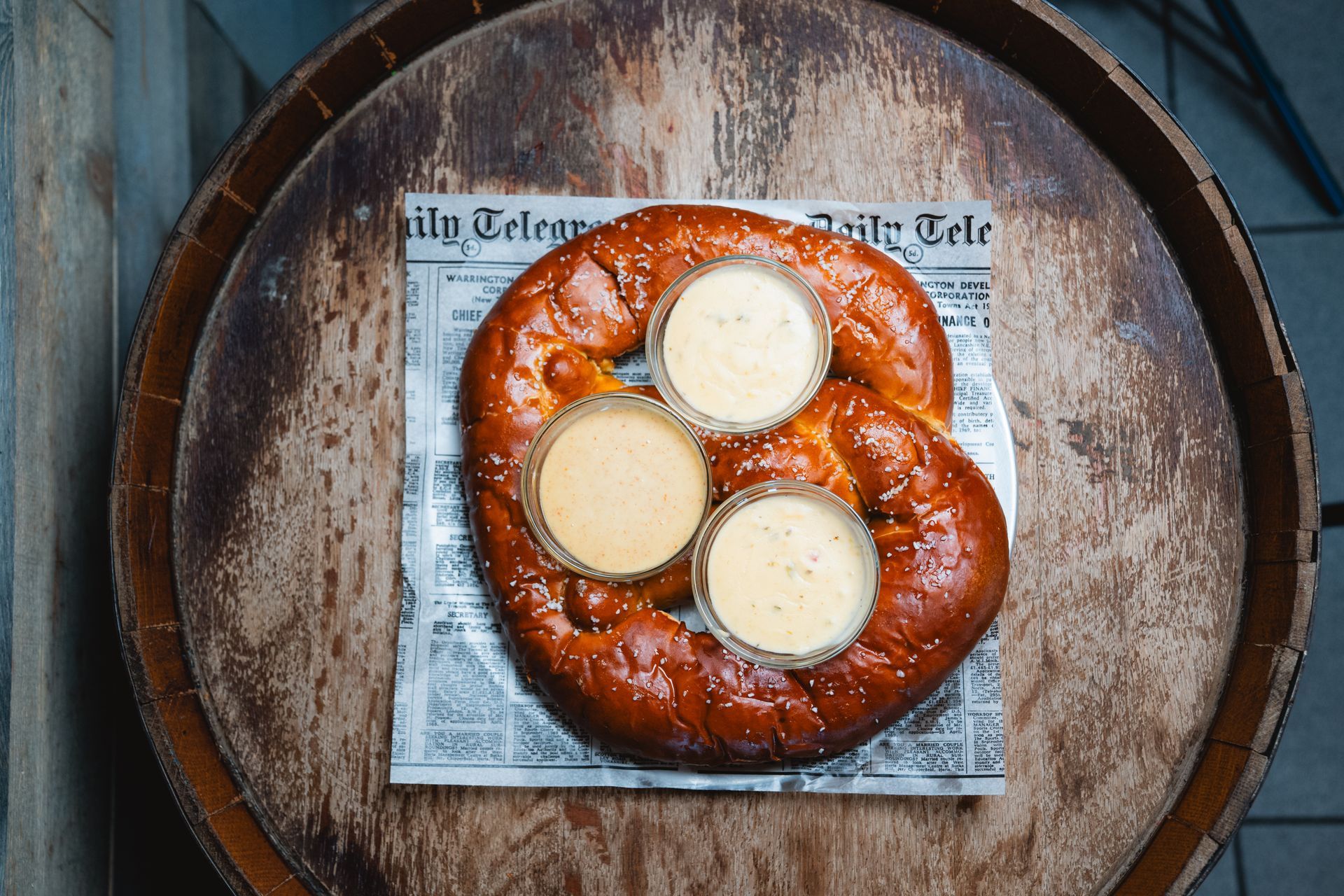 Giant pretzel with three dipping sauces, served on newspaper on a wooden barrel.