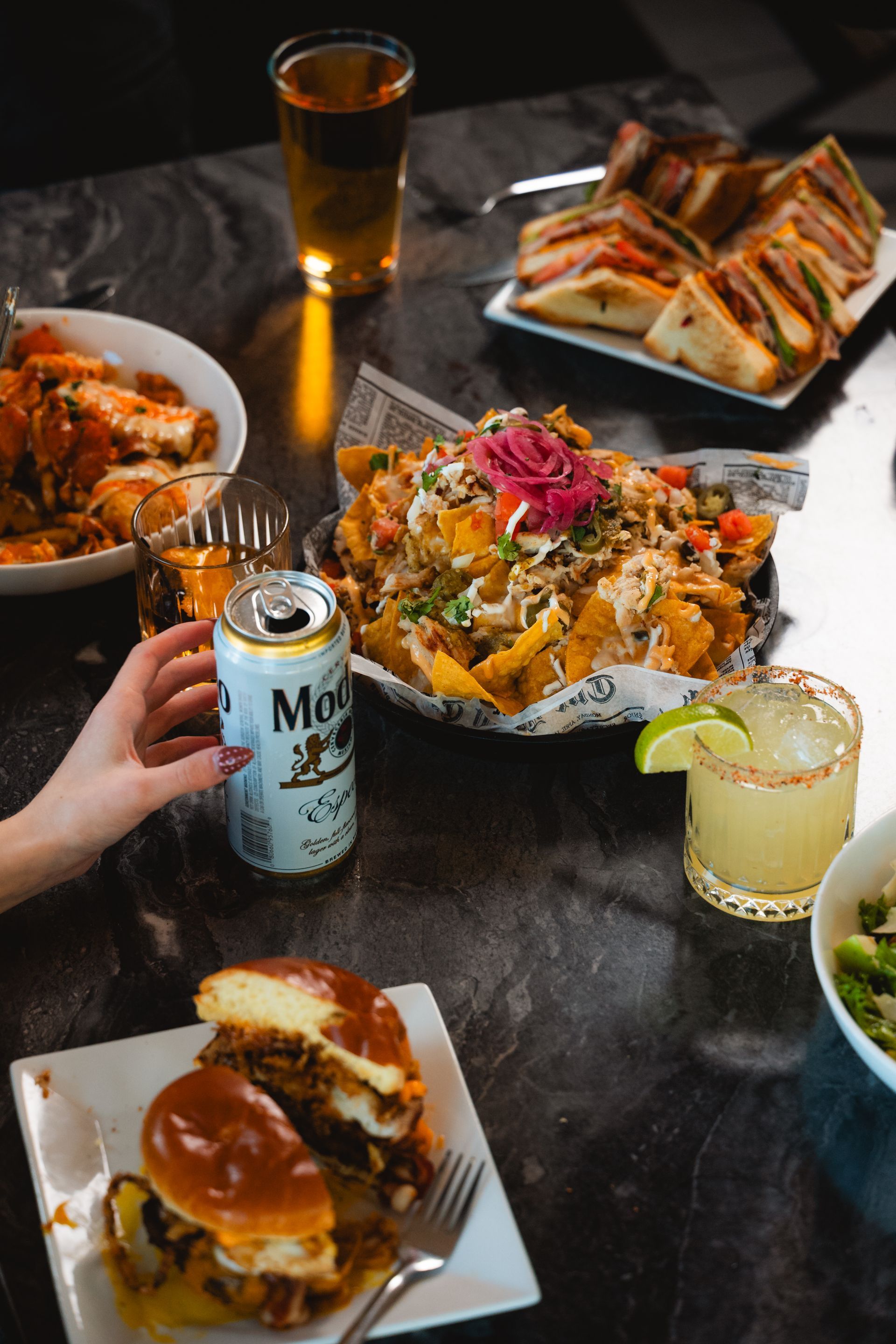 Table of various bar foods with drinks: nachos, sandwiches, sliders, wings, margarita, beer can, and a hand.