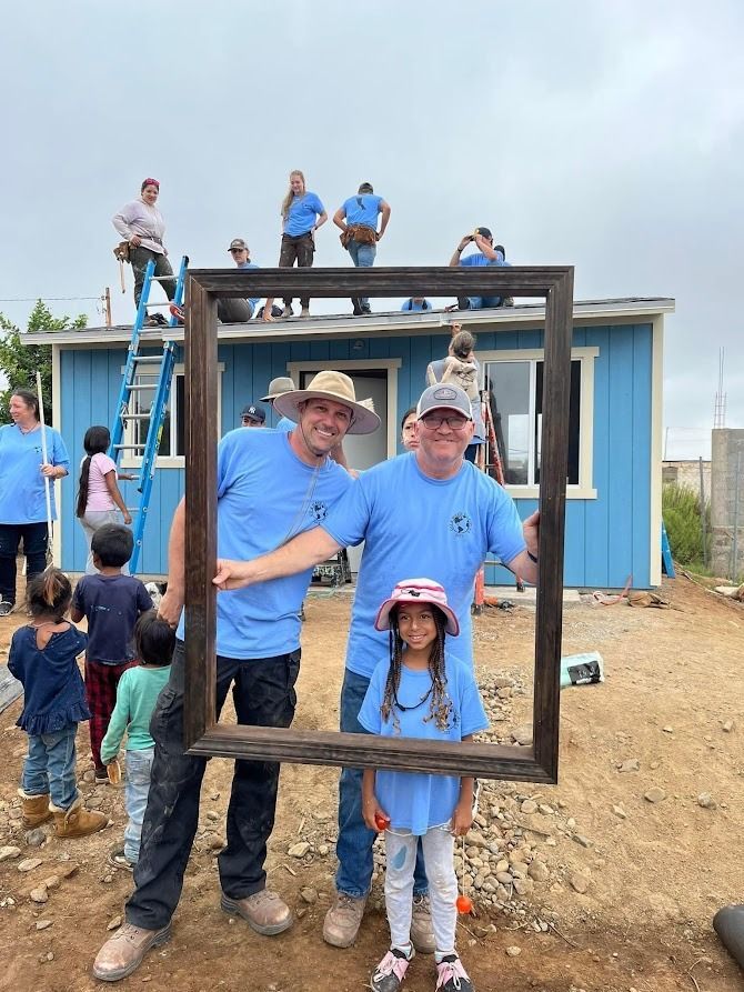 A group of people are standing in front of a blue house holding a picture frame.