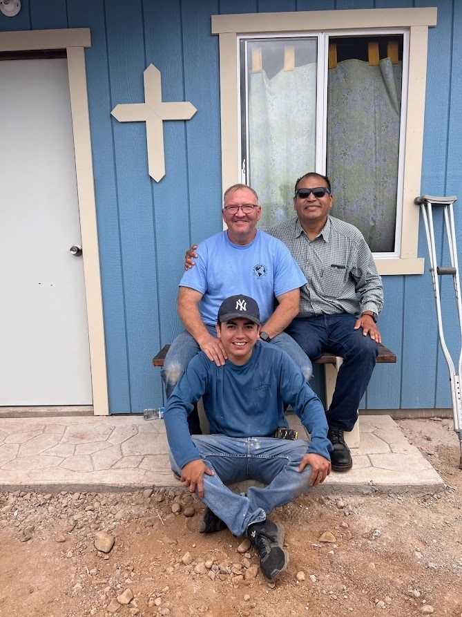 Three men are posing for a picture in front of a blue house with a cross on it