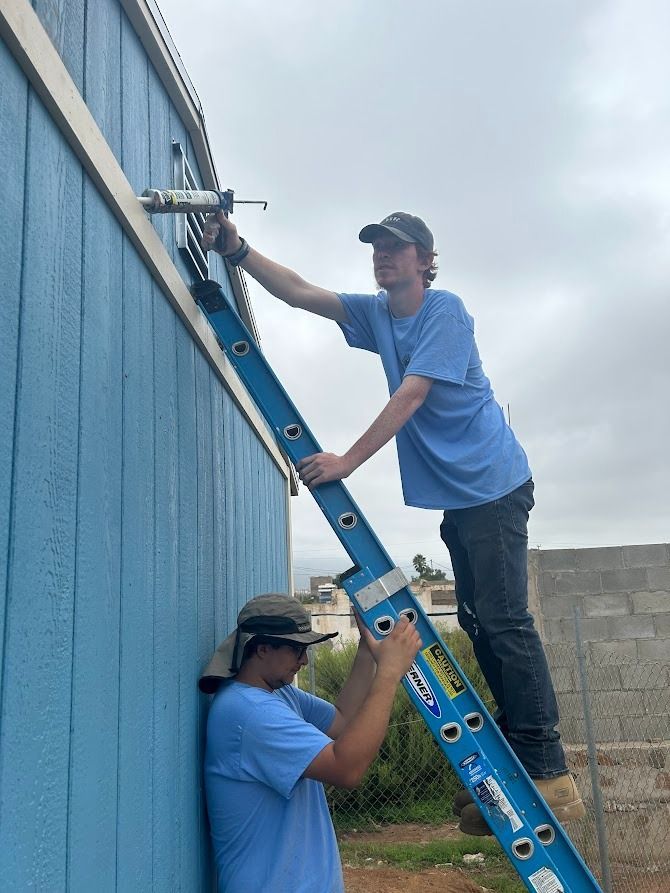 Two men are working on a blue building with a ladder.