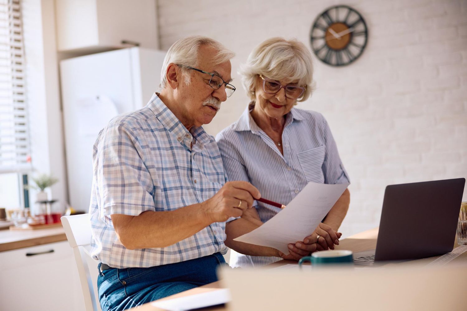 Two people look at a document together at a table with a laptop, focusing on their shared work in a bright room.