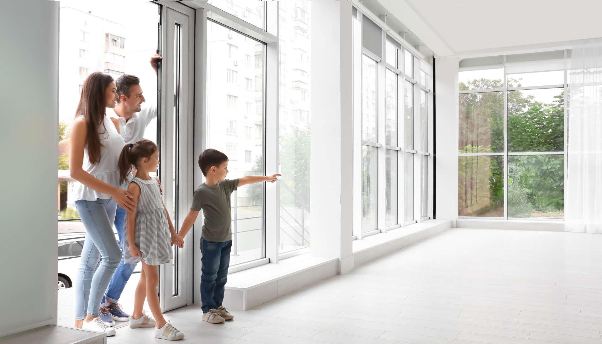 A family stands inside a bright, empty room with large windows, looking out and pointing at the view.