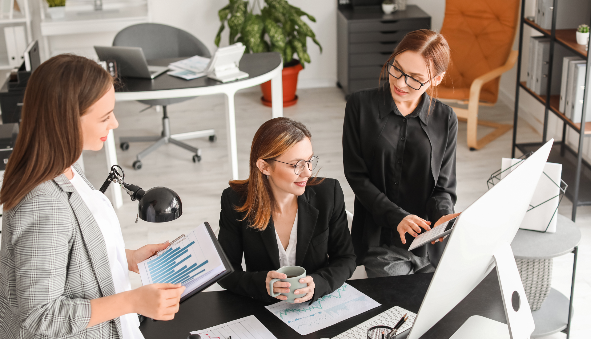 Three professionals in a modern office collaborate around a desk, reviewing data charts and a computer screen.