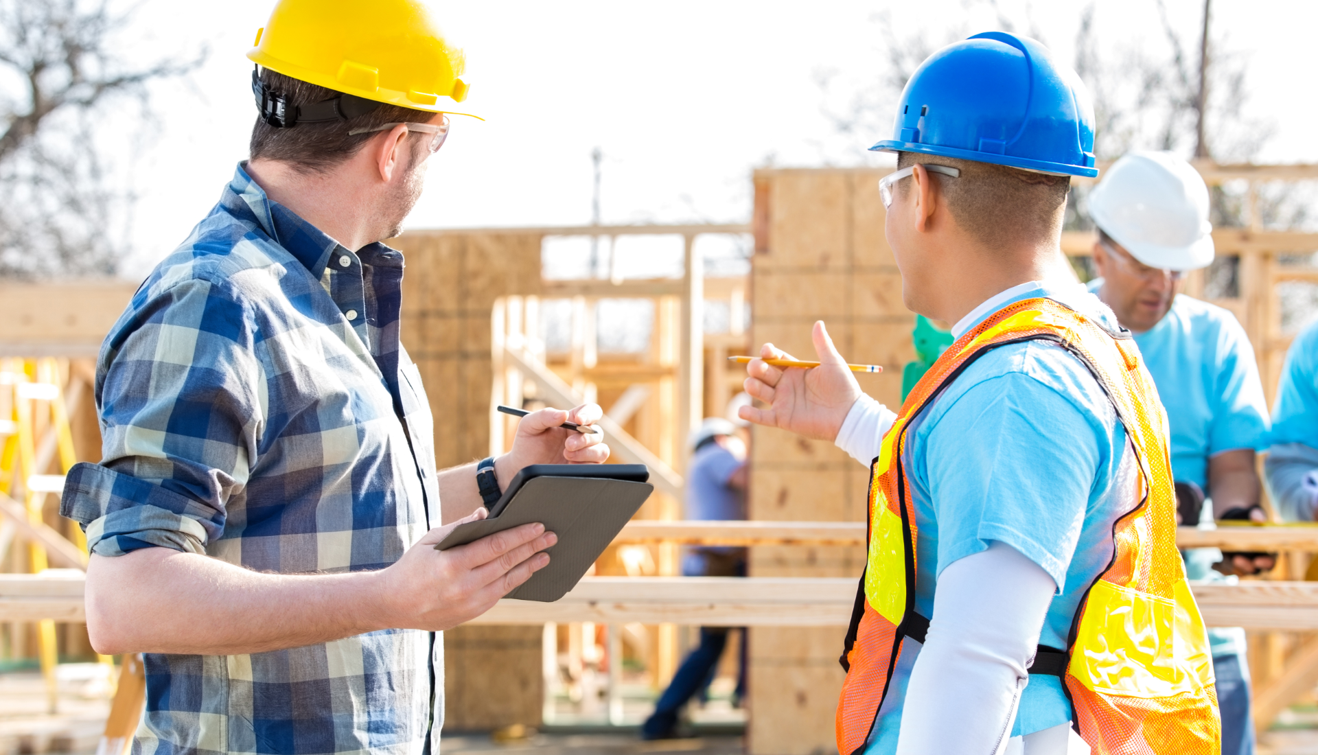 Two construction workers in hard hats and vests discuss plans on a tablet at an outdoor timber framing job site.