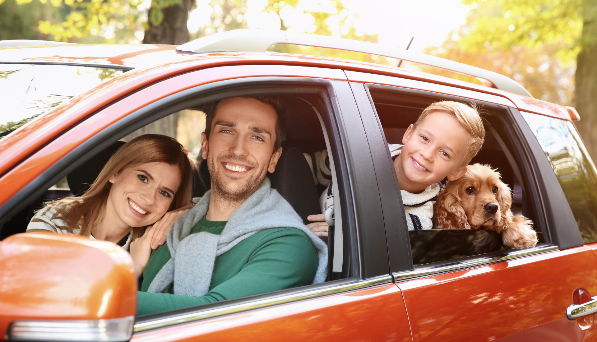 A family and their dog smiling from inside an orange car on a sunny day.