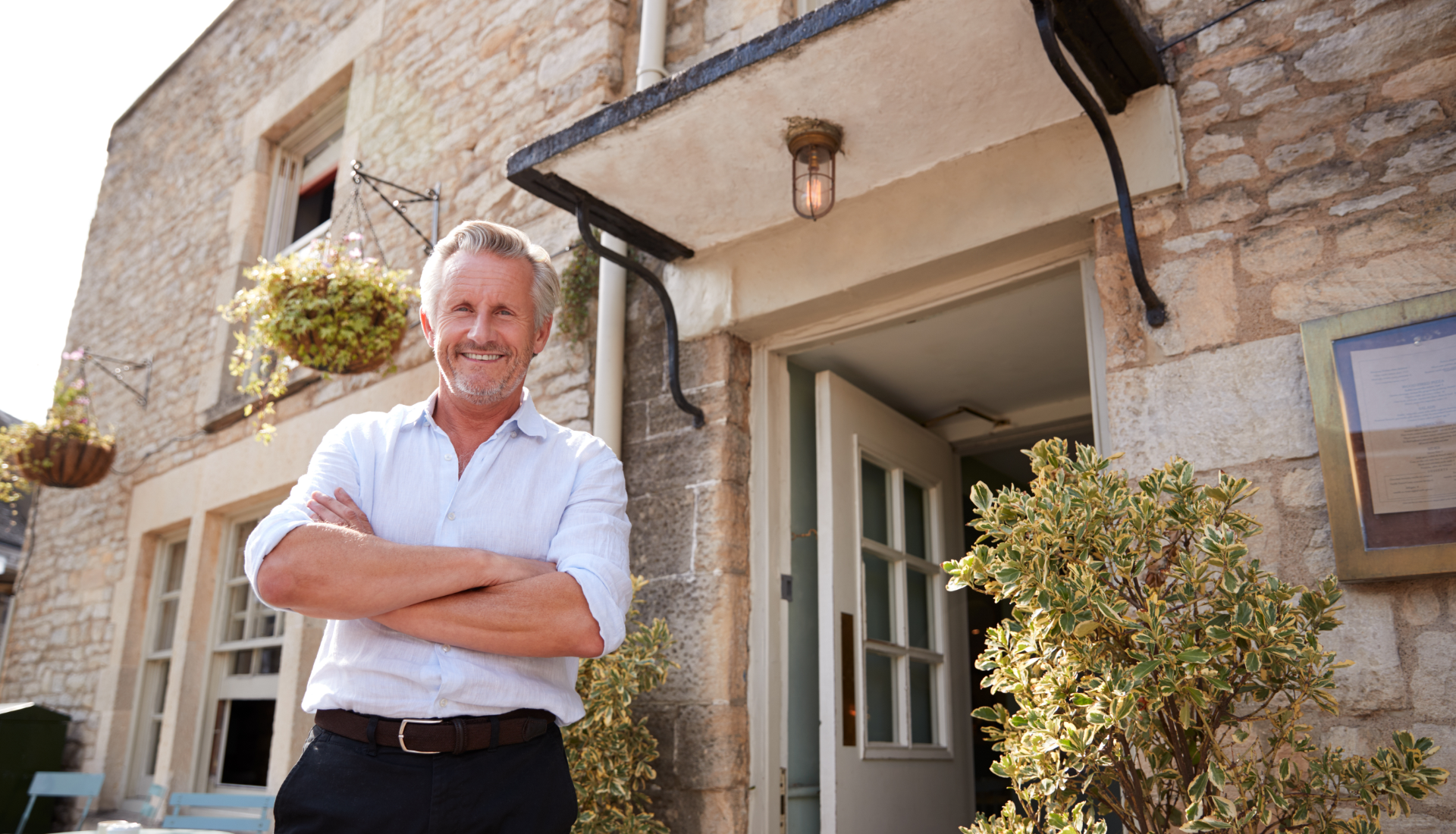 A smiling man in a light shirt stands with arms crossed in front of a stone building entrance.