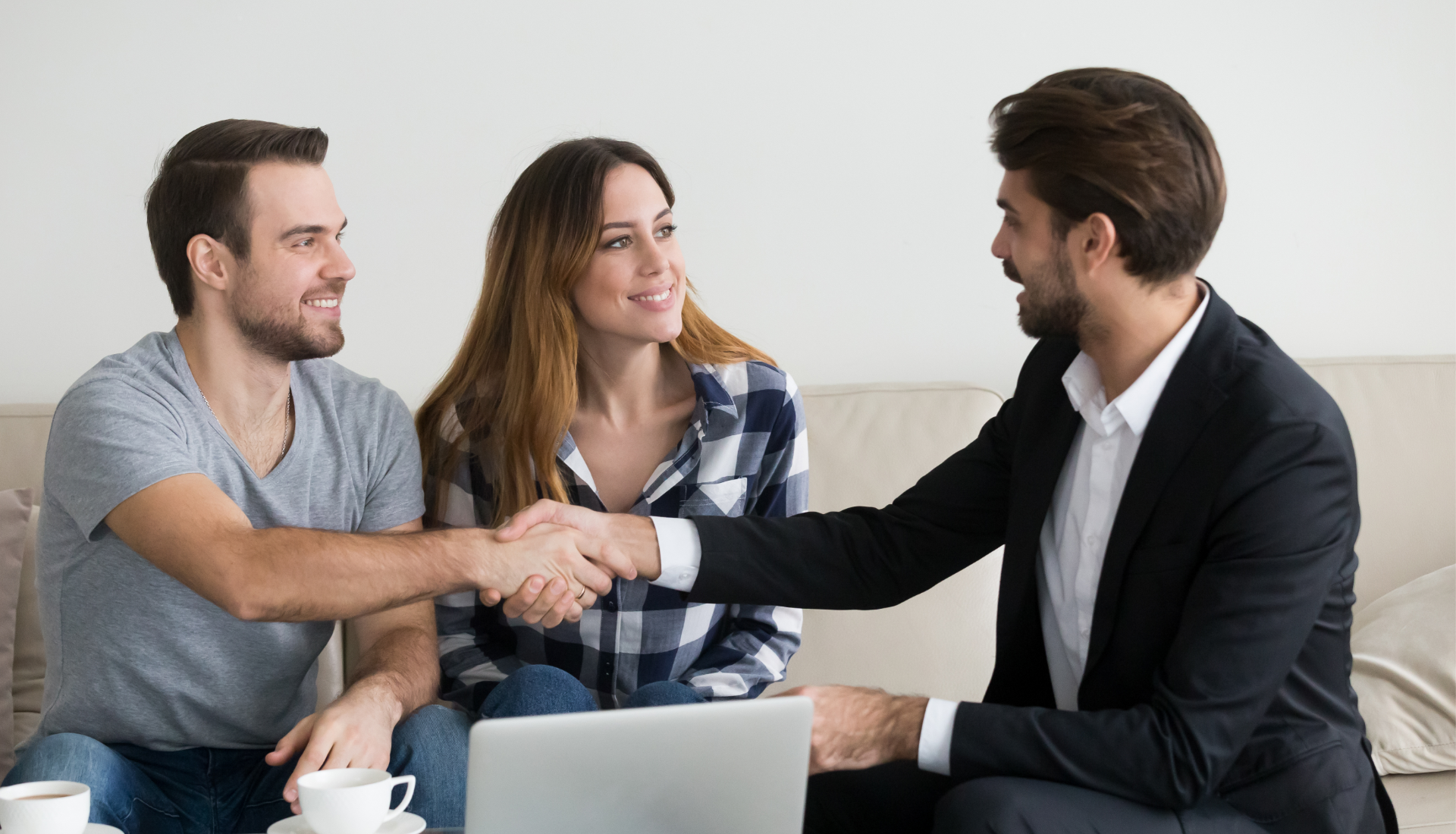 A person in a business suit shakes hands with another person, while a third person watches, seated on a sofa with a laptop.