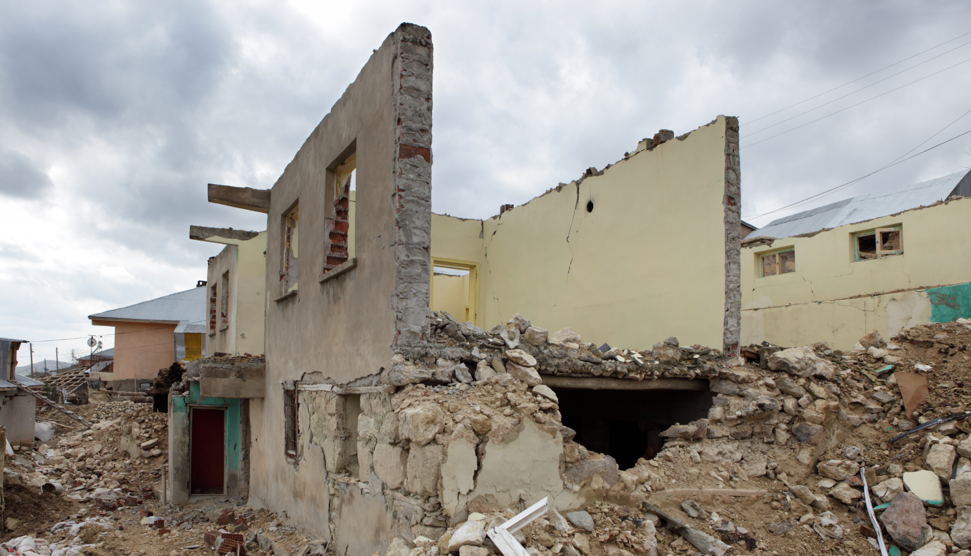 A partially destroyed two-story beige house surrounded by rubble under a cloudy sky.