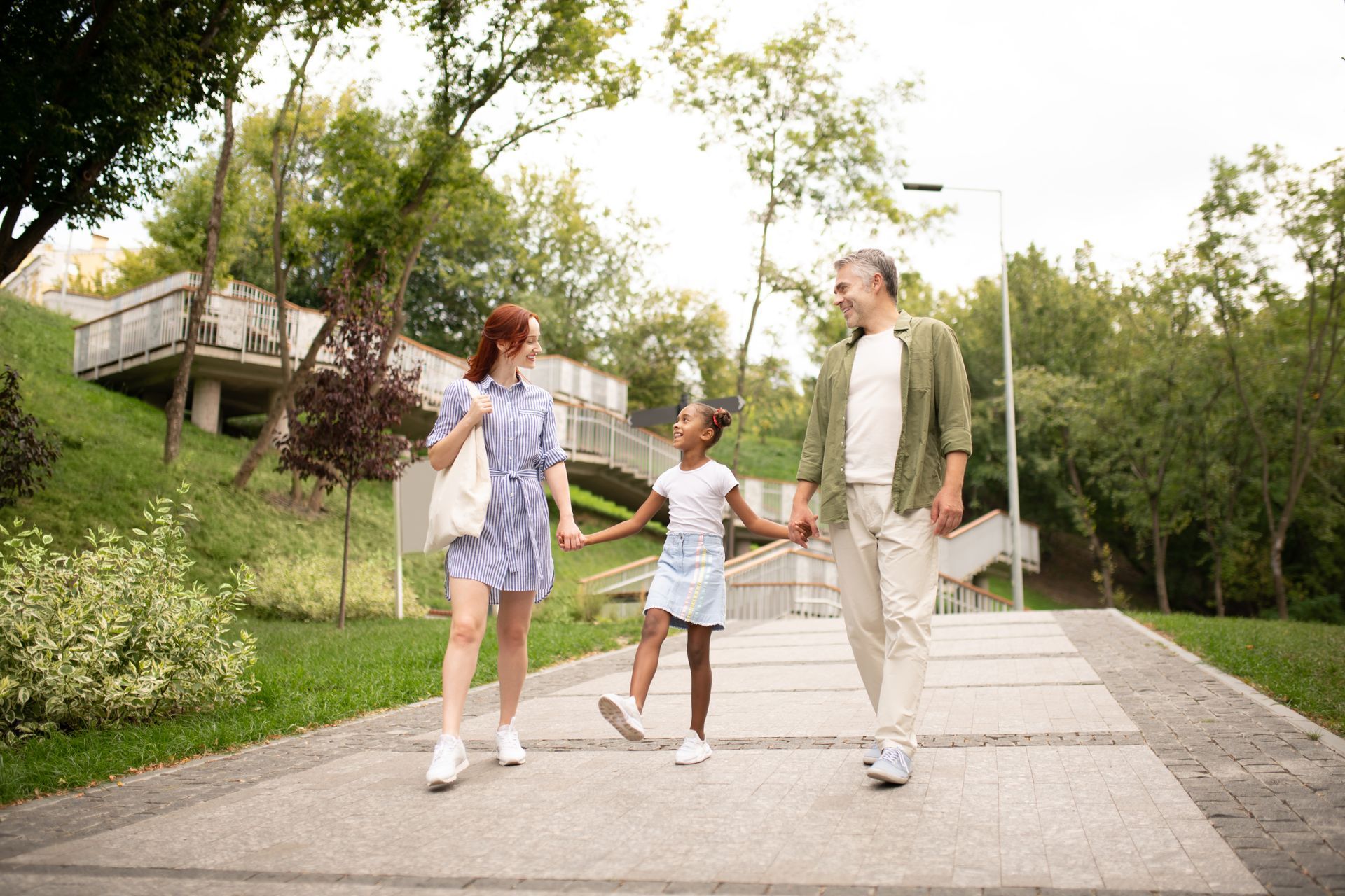 A family walks together on a park path, holding hands and smiling on a sunny day.