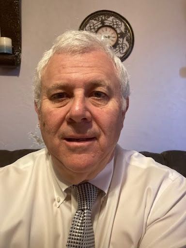 A person with short white hair wearing a white collared shirt and a patterned tie, posing for a portrait indoors.