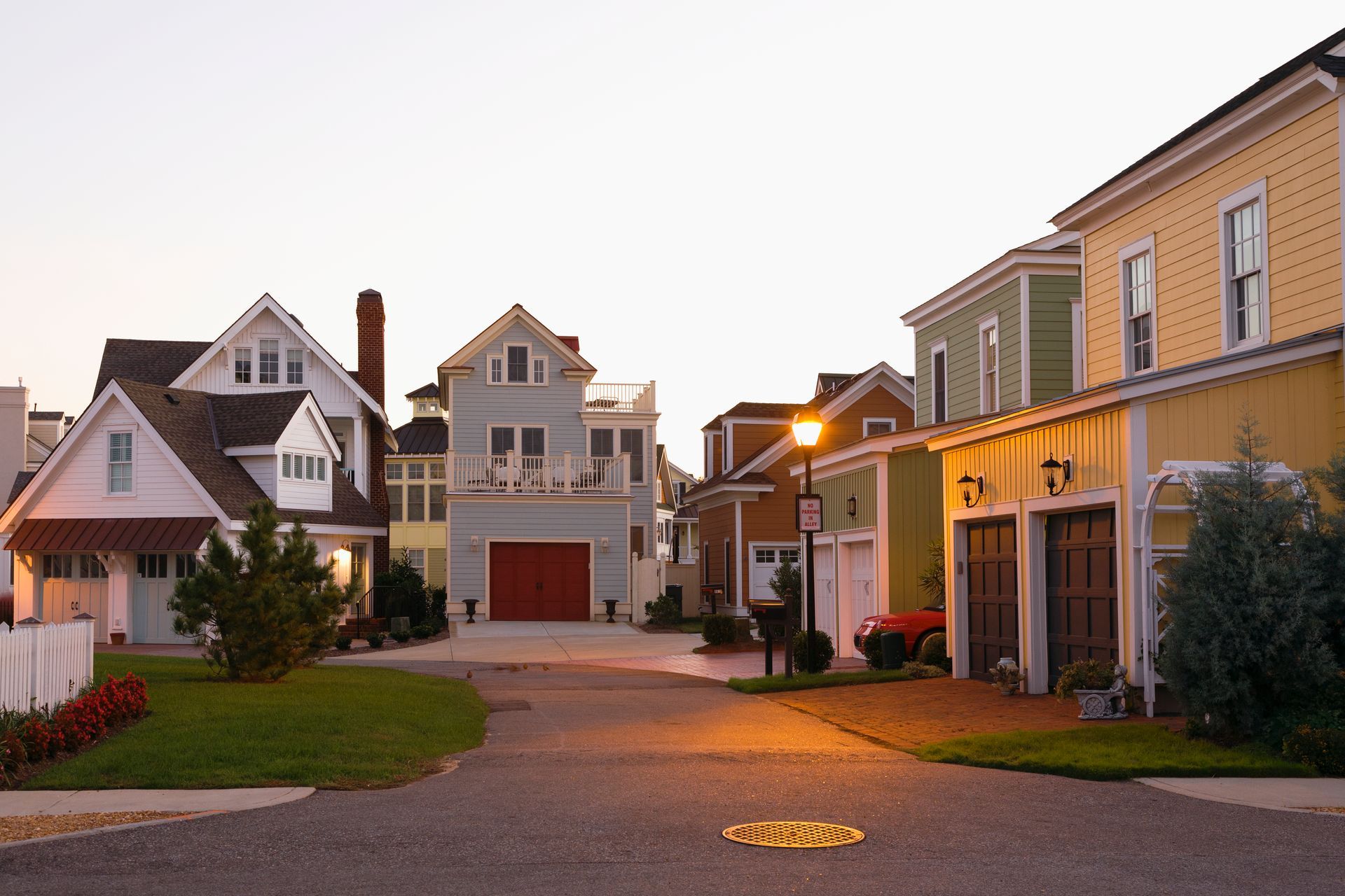 A quiet residential street at dusk, lined with colorful houses, a glowing streetlamp, and paved driveways.
