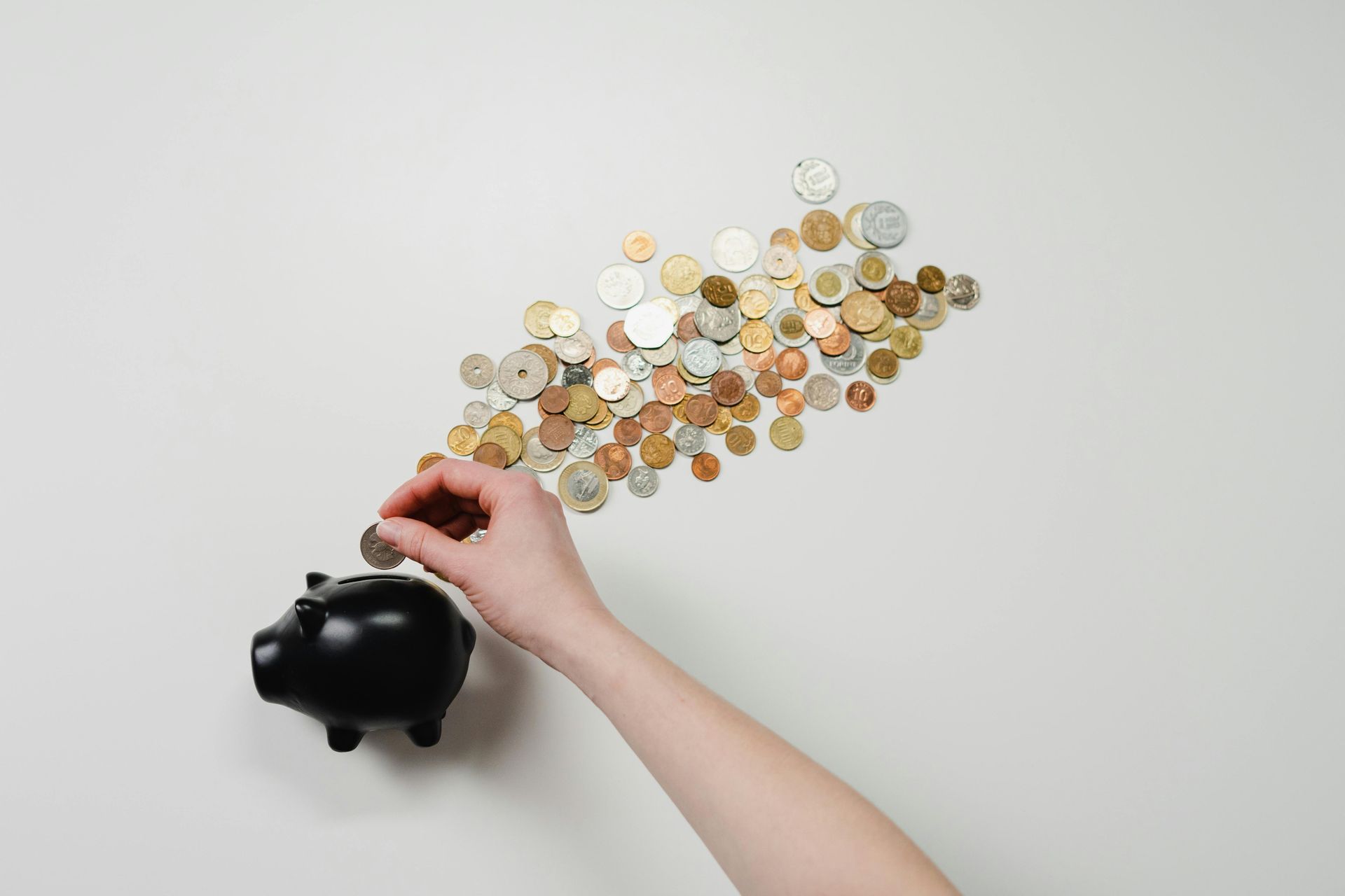 Hand putting a coin into a black piggy bank, coins scattered in the air on a white background.