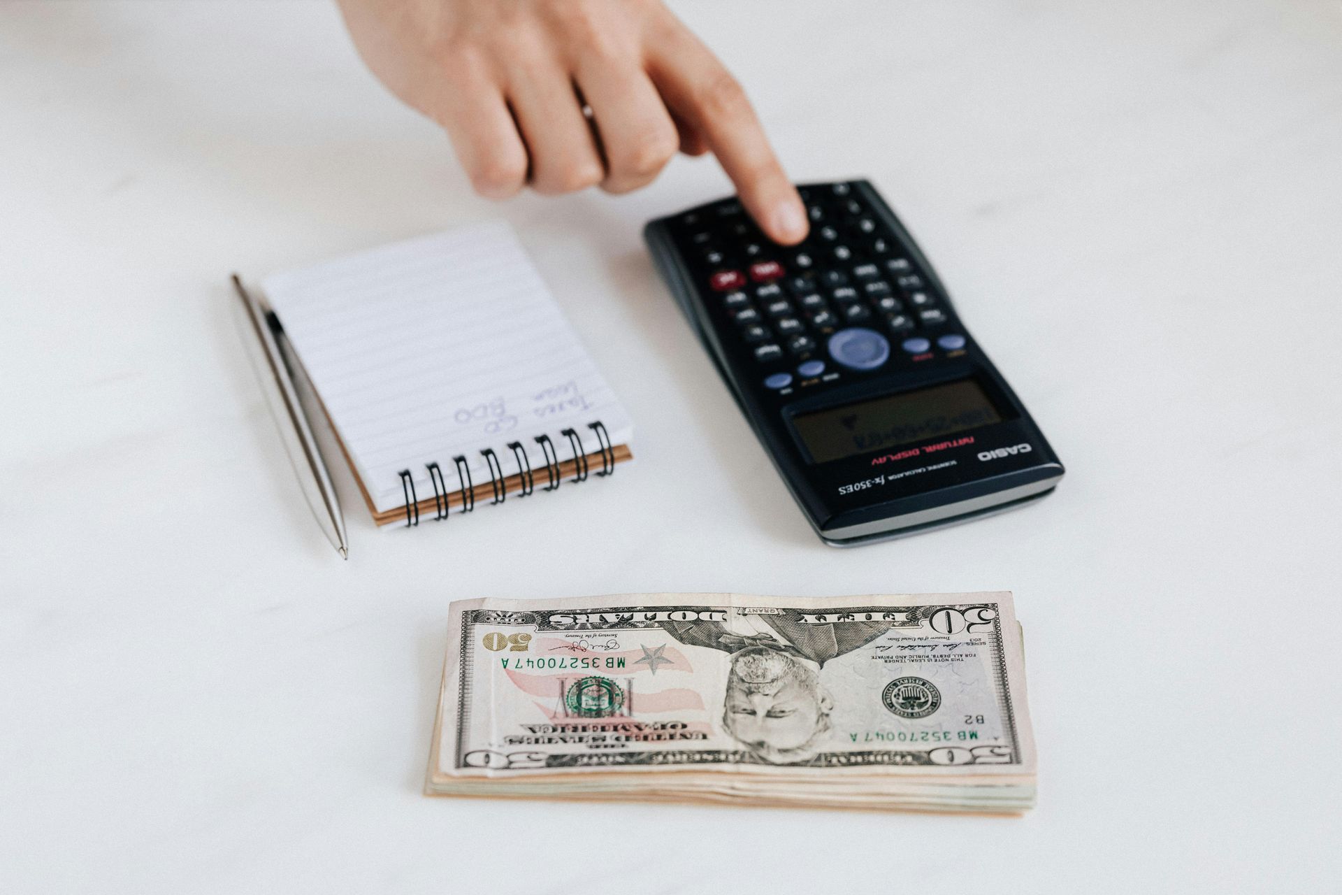 Hand using a calculator, next to a notepad with a pen and a stack of cash on a white surface.