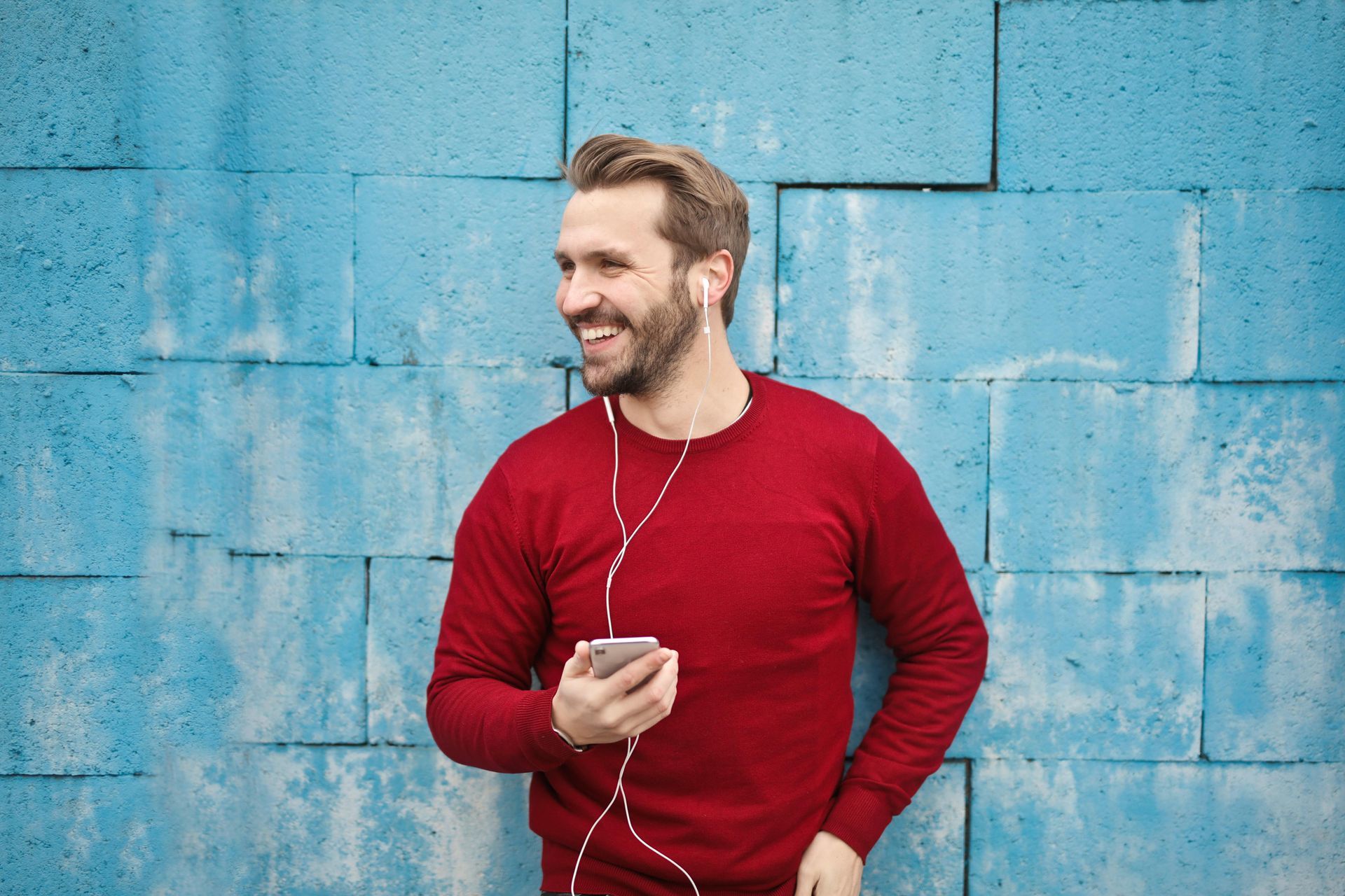 Man in red sweater smiles, listening to music with wired earphones, holding phone near blue brick wall.