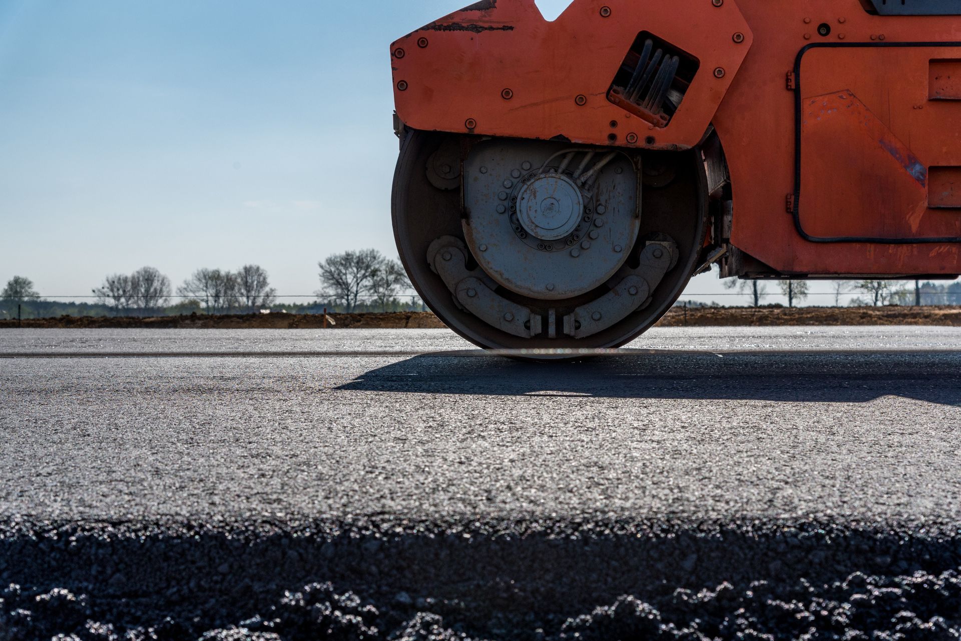 Close view of an orange road roller as it flattens an asphalt road, with the sky as background