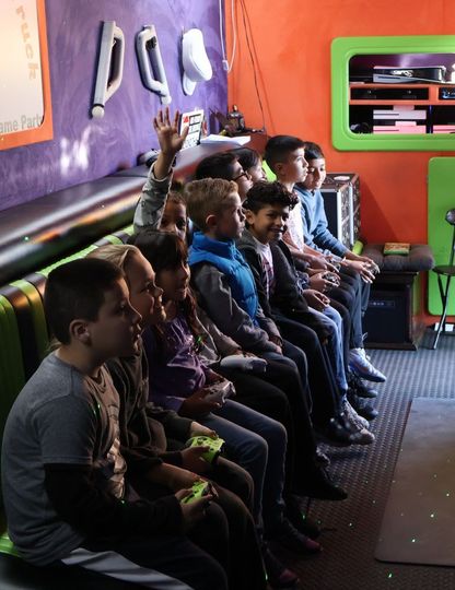 Children seated, some with controllers, in a brightly colored gaming room. One raises their hand.