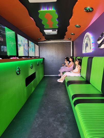 Inside a brightly colored gaming truck: Three girls seated on a green bench, playing video games on multiple screens.