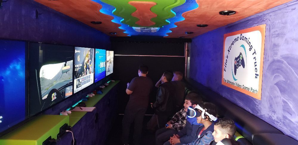 Inside a game truck: people watch screens playing video games. Colorful ceiling and logo on the wall.