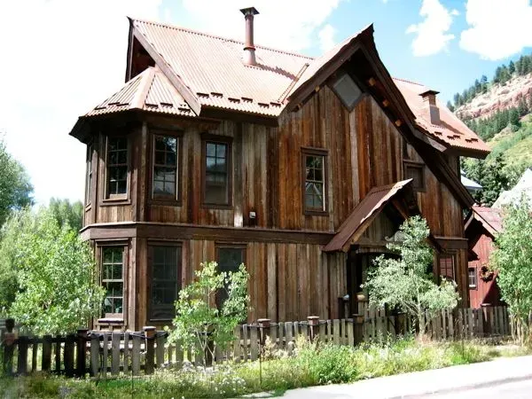 Weathered wooden house with metal roof, small fence, and trees.